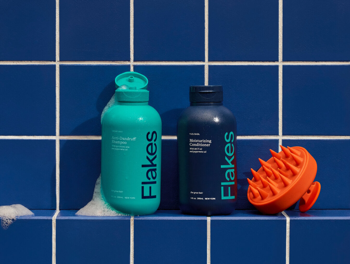 Turquoise shampoo and navy conditioner bottles with an orange scalp massager on blue tiled background.